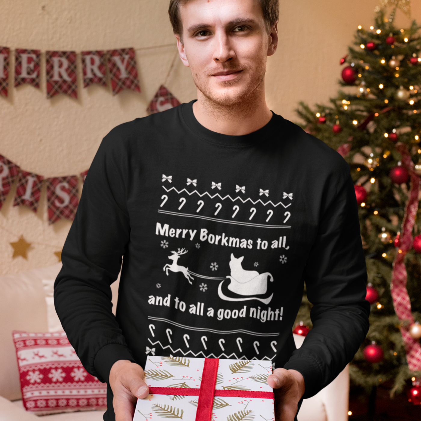 A model wearing a black long sleeved shirt with the text "Merry Borkmas to all, and to all a good night!" with a corgi in Santa's sled. He's in front of a room with Christmas decorations and he's holding a present.