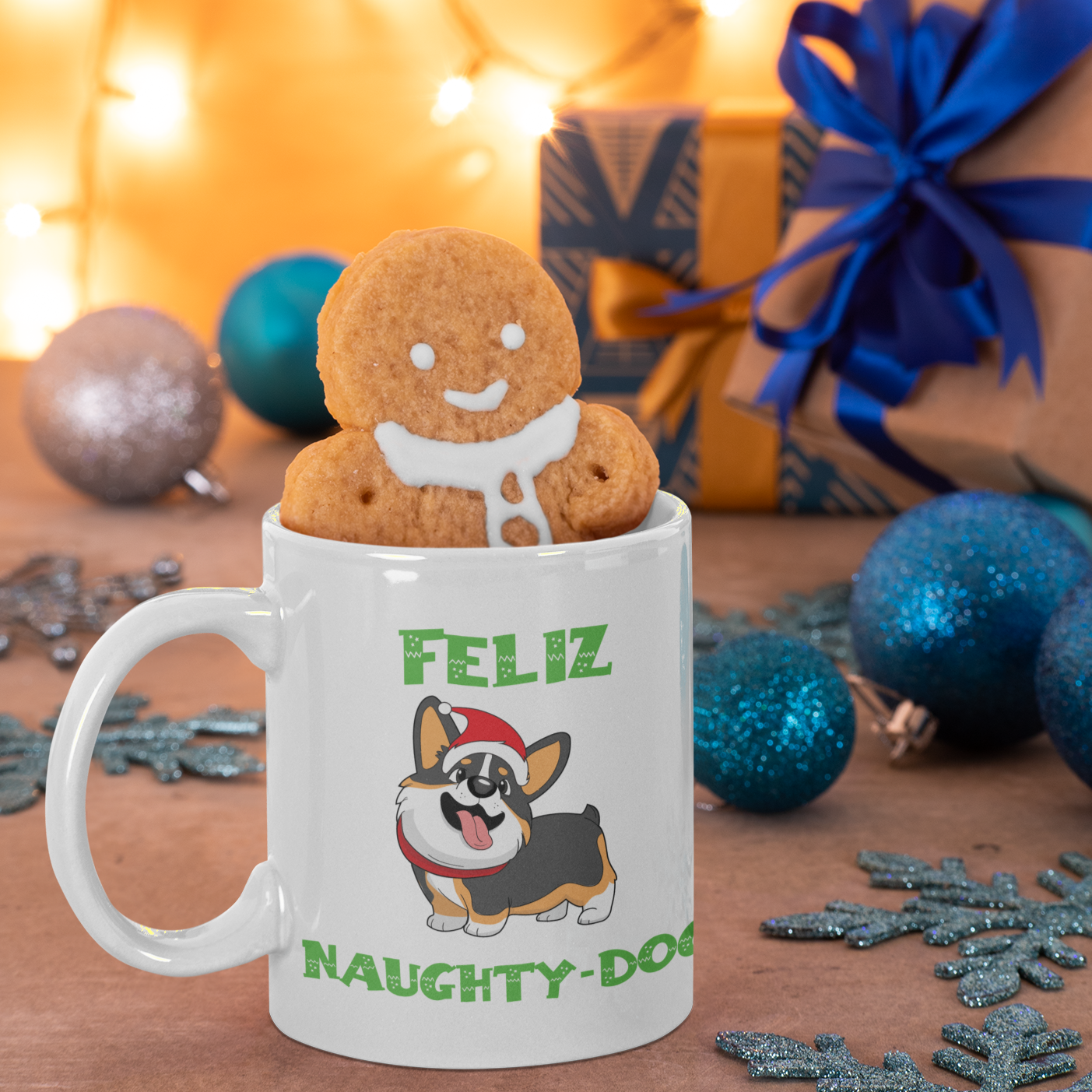 A mug that says "Feliz Naught-Dog" and has a smiling tricolor corgi wearing a Santa hat on it. The mug has a gingerbread cookie in it and is amongst Christmas decor and presents.