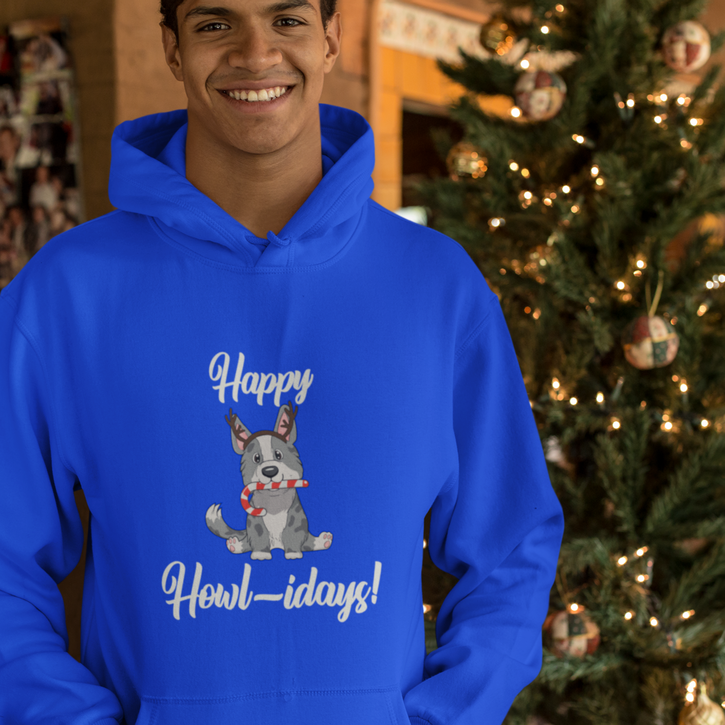 A model is wearing a blue hoodie that has a cardigan corgi on it. The corgi is wearing fake reindeer horns and is holding a candy cane in its mouth. The text says "Happy Howl-idays". The model is standing in front of a Christmas tree.