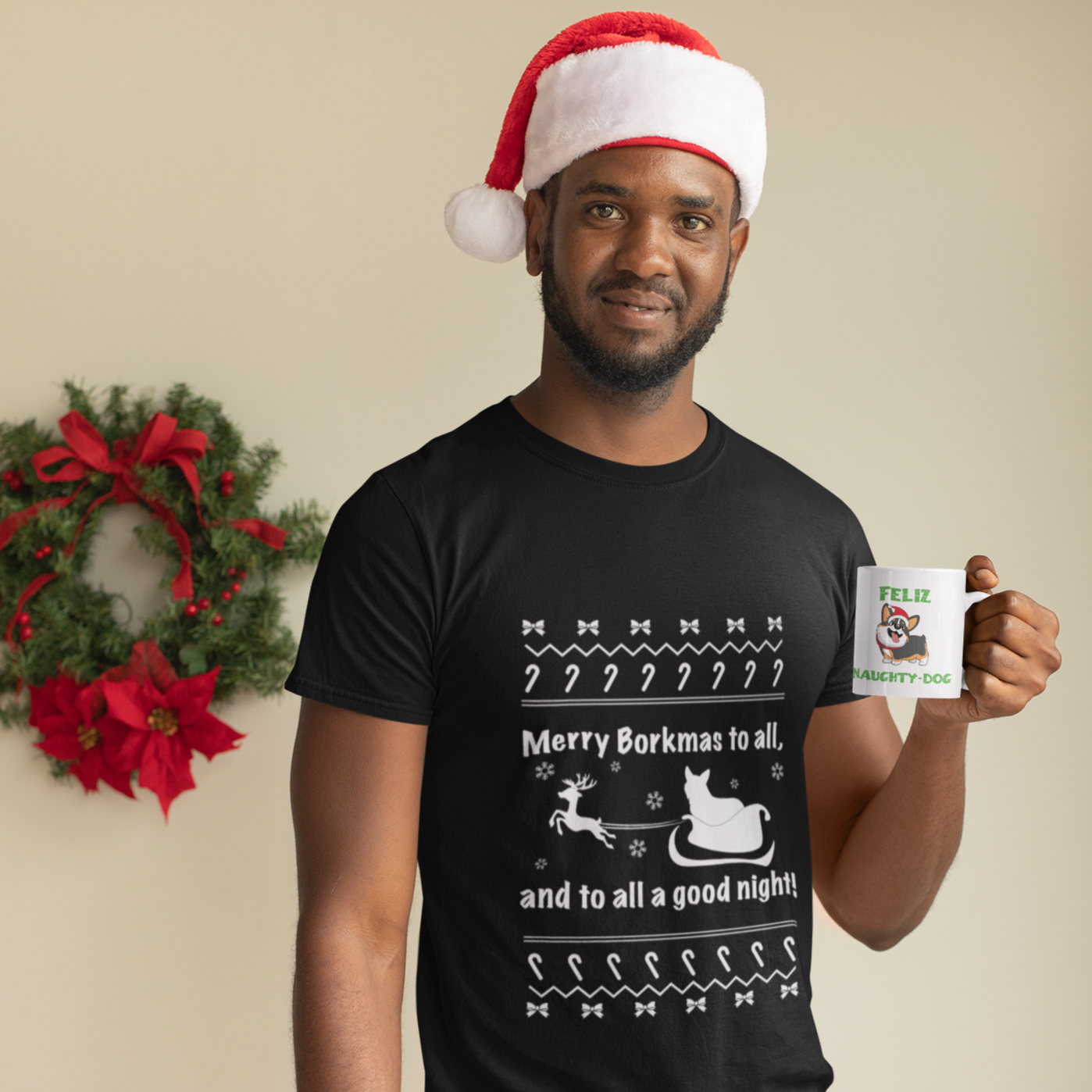A model wearing a black, short sleeved shirt that says "Merry Borkmas to all, and to all a good night!" with a Christmas pattern, and a corgi in Santa's sleigh. The model is standing in front of a wreath and he's wearing a Santa hat. He's holding a mug with our Feliz Naught-Dog design on it.