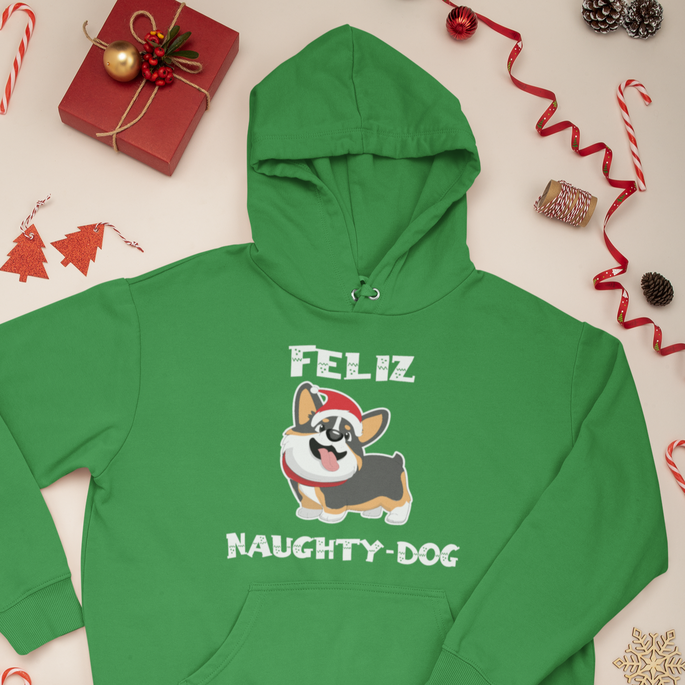 A green hoodie that says "Feliz Naught-Dog" and has a smiling tricolor corgi wearing a Santa hat on it. The hoodie is laying flat amongst Christmas decor and presents.
