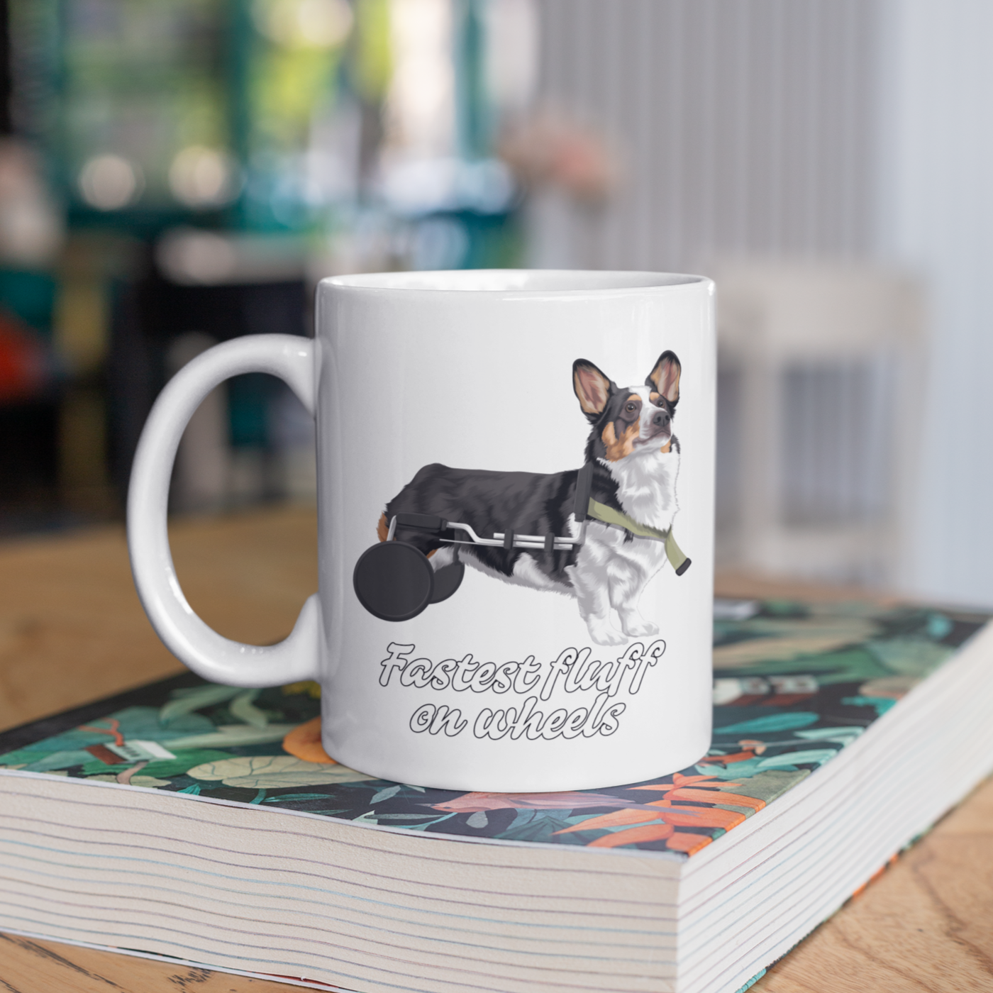 A white coffee mug sitting on a book on a table. The design on the mug is a tricolor corgi that is using a dog wheelchair. There is black-outlined white text that says "Fastest fluff on wheels".