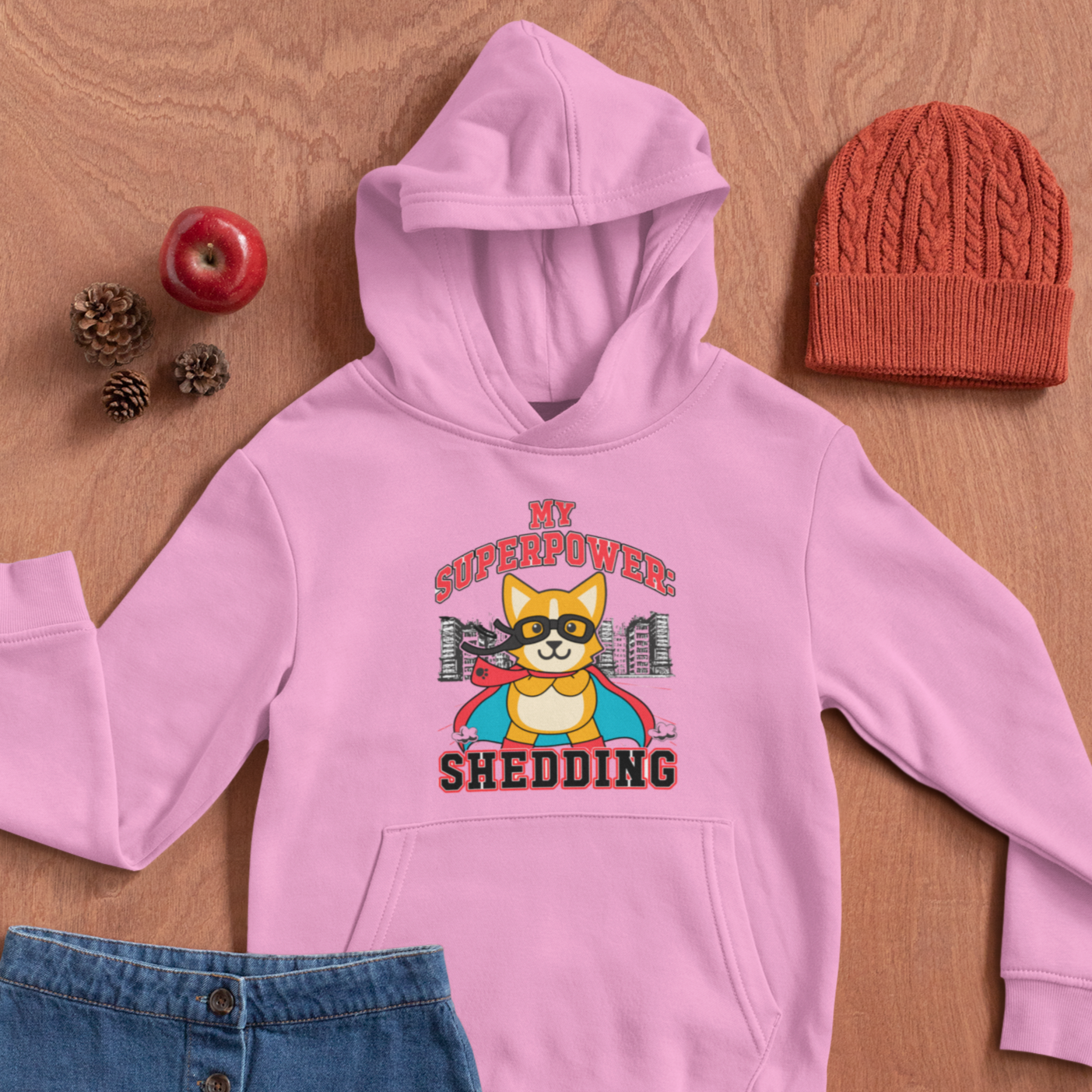 A pink hooded sweatshirt laying on a table next to a knit hat, some pinecones, an apple, and a red apple. The hoodie is pink and has red and black text that says "My superpower: Shedding" with a corgi dressed as a superhero. 