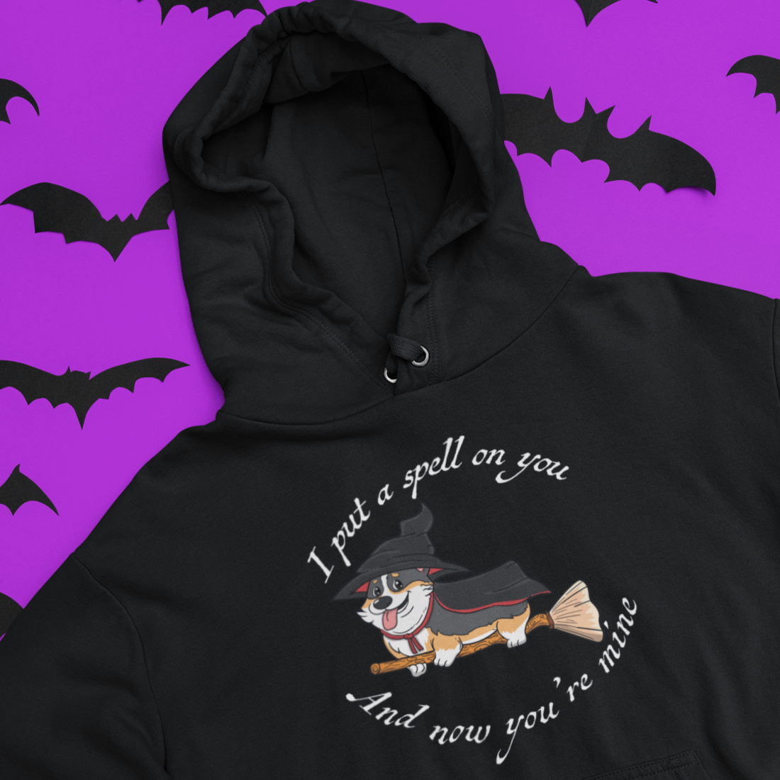 A black hooded sweatshirt laying on a purple table. There are black paper bats also on the table. The design on the hoodie is a tricolor corgi dressed up as a witch and riding a broom. The text "I put a spell on you and now you're mine" go in a circle above and below the corgi. 