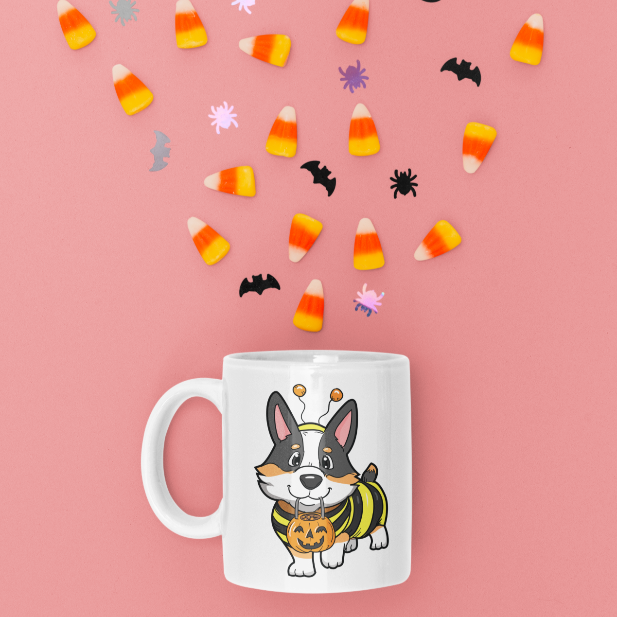 A white coffee mug surrounded by halloween candy. The design on the mug is a tricolor corgi wearing a bumblebee costume.