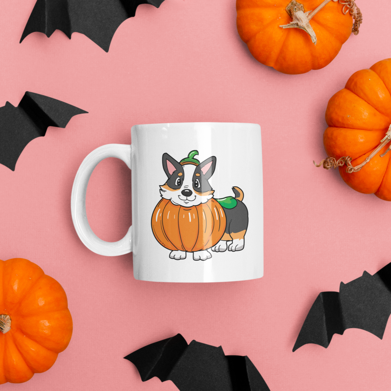 A white coffee mug is laying on a table next to some paper bat decorations and some miniature pumpkins. The design on the mug is a tricolor corgi dressed up as a pumpkin. 