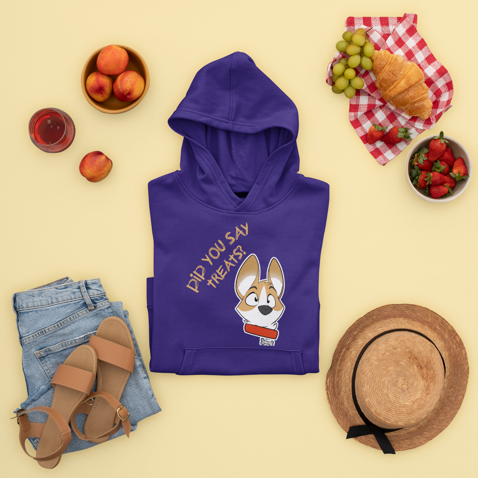 A folded hooded sweatshirt on a table with an assortment of food and in the bottom corners, clothing. The hooded sweatshirt is dark purple and depicts a surprised looking corgi. The text says "Did you say treats?"