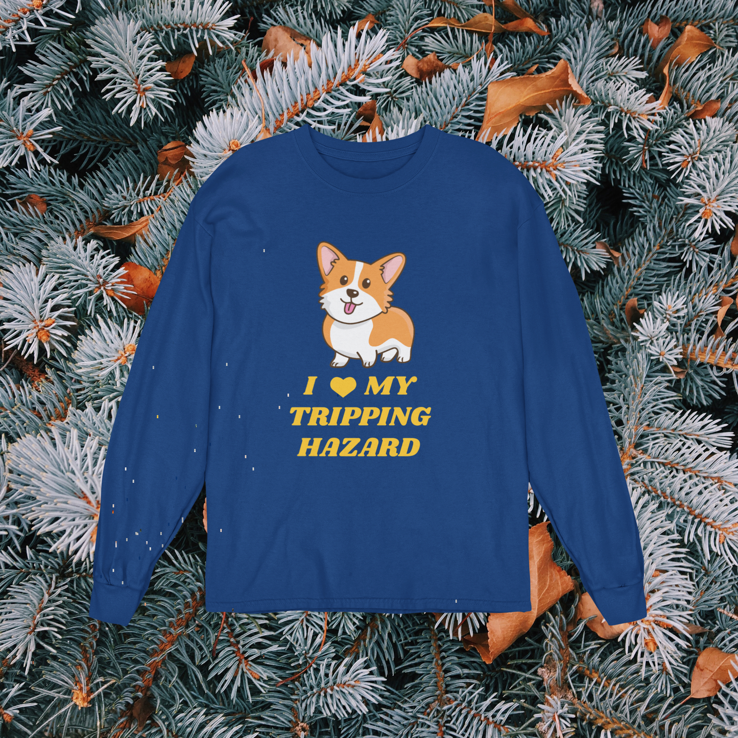 A dark blue, long sleeved t shirt on a background of evergreen tree branches. The shirt has a corgi with its tongue sticking out and yellow text that says "I love my tripping hazard". 