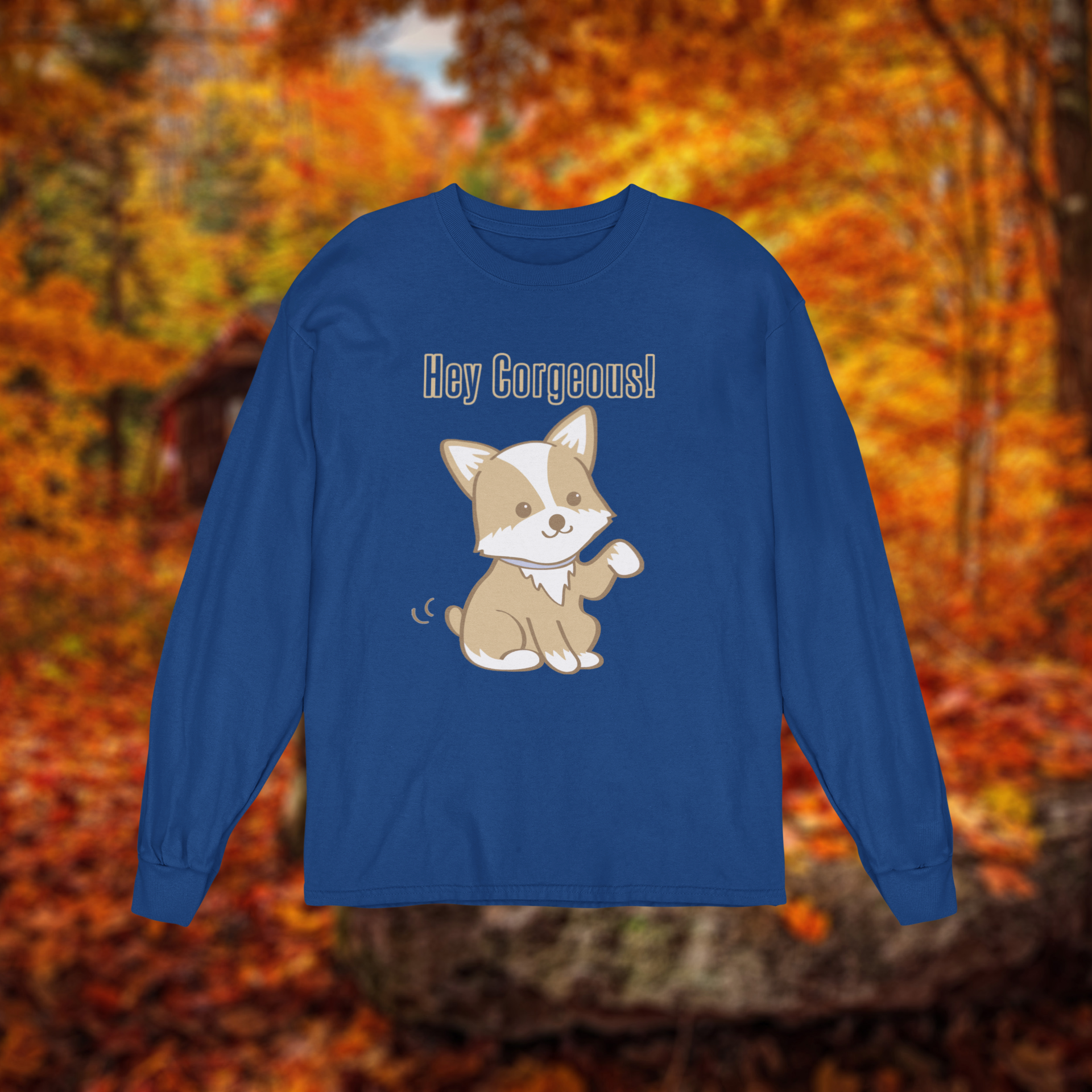 A dark blue long sleeved t shirt on a background of a fall forest. The shirt ha a corgi waiving it's paw and it says "Hey Corgeous" in yellow text. 