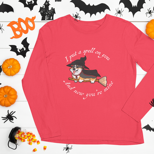 A flat lay of a long sleeved t shirt. The shirt is red and has white text that says: I put a spell on you and now you're mine. There is also a tricolor corgi dressed as a witch while riding a broom. The shirt is surrounded by paper halloween decor. 