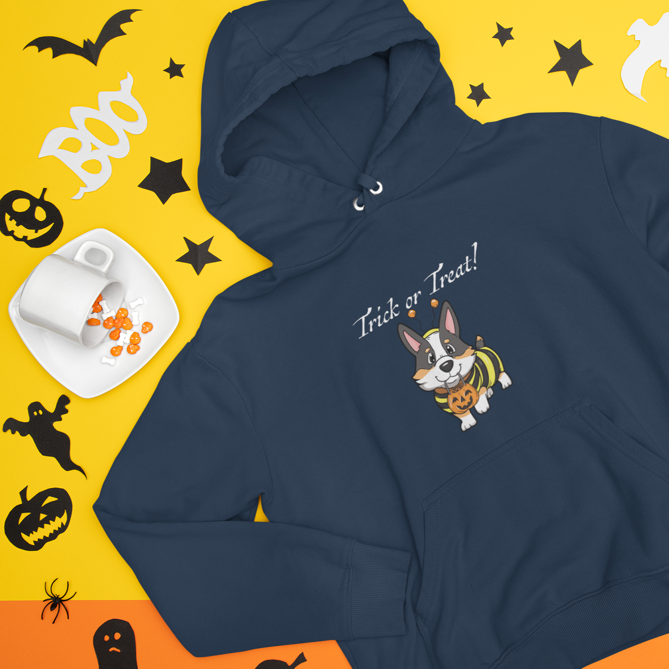 A flat lay of a dark blue hooded sweatshirt. The hoodie is among some halloween decorations and halloween candy. The design on the hoodie is a tricolor corgi dressed up in a halloween costume. The text on the hoodie is "Trick or Treat!"