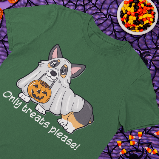 A green t shirt laying on a purple table. Next to the shirt are some halloween decorations and halloween candy. The green shirt has a tricolor corgi dressed up as a ghost. The corgi is holding a halloween pumpkin in it's mouth and the text under it says "Only treats please!".