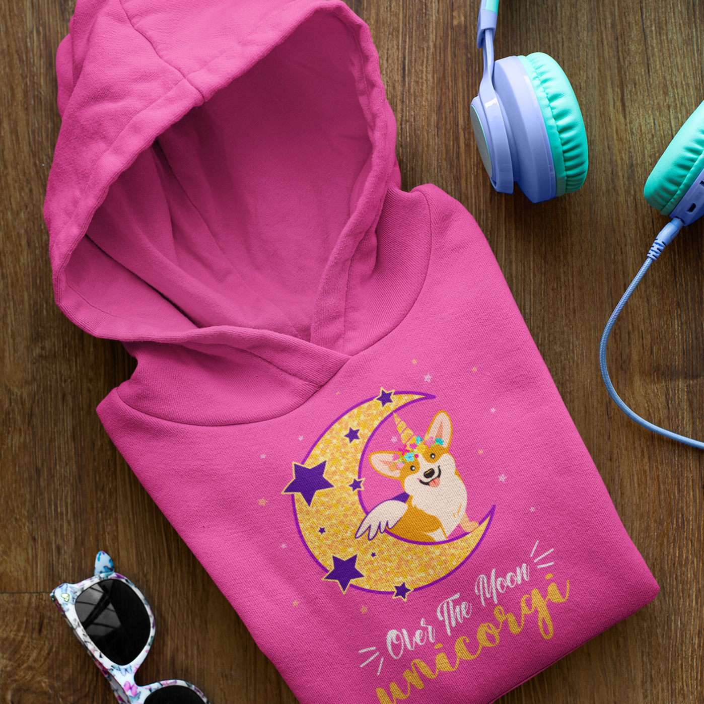 A folded hooded sweatshirt on a table next to a pair of sunglasses. The pink hoodie has a corgi that has a unicorn and wings while sitting on a crescent moon. The text says "Over the moon Unicorgi".