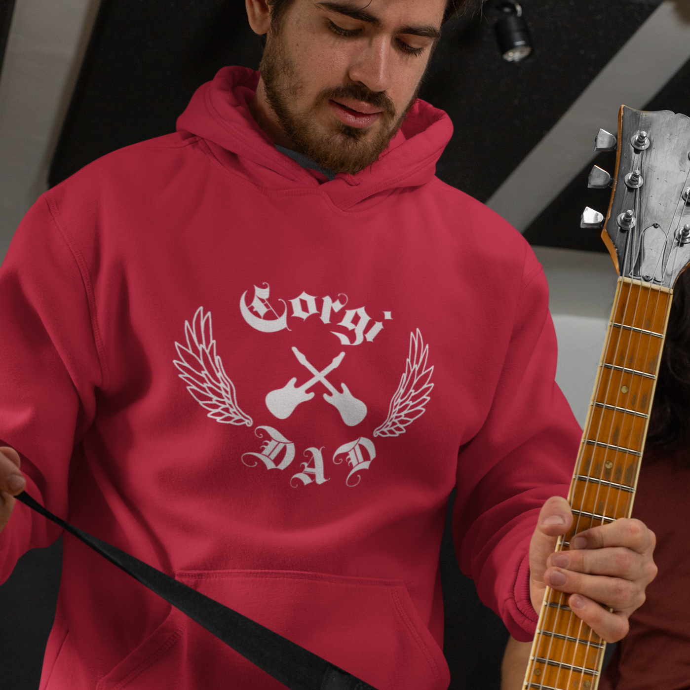 A man wearing a red hooded sweatshirt. The design on the hoodie is crossed electric guitars, lined wings, and "Corgi Dad" in old english font. The man is also holding an electric guitar. 