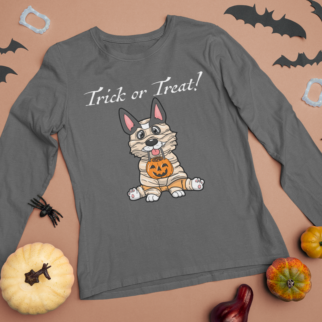 A flat lay of a dark grey, long sleeved t shirt. The design of the shirt is a tricolor corgi dressed up as a mummy, and is holding a pumpkin trick or treat container. The text above the dog is white and says "Trick or Treat!". There are halloween decorations on the table near the shirt. 