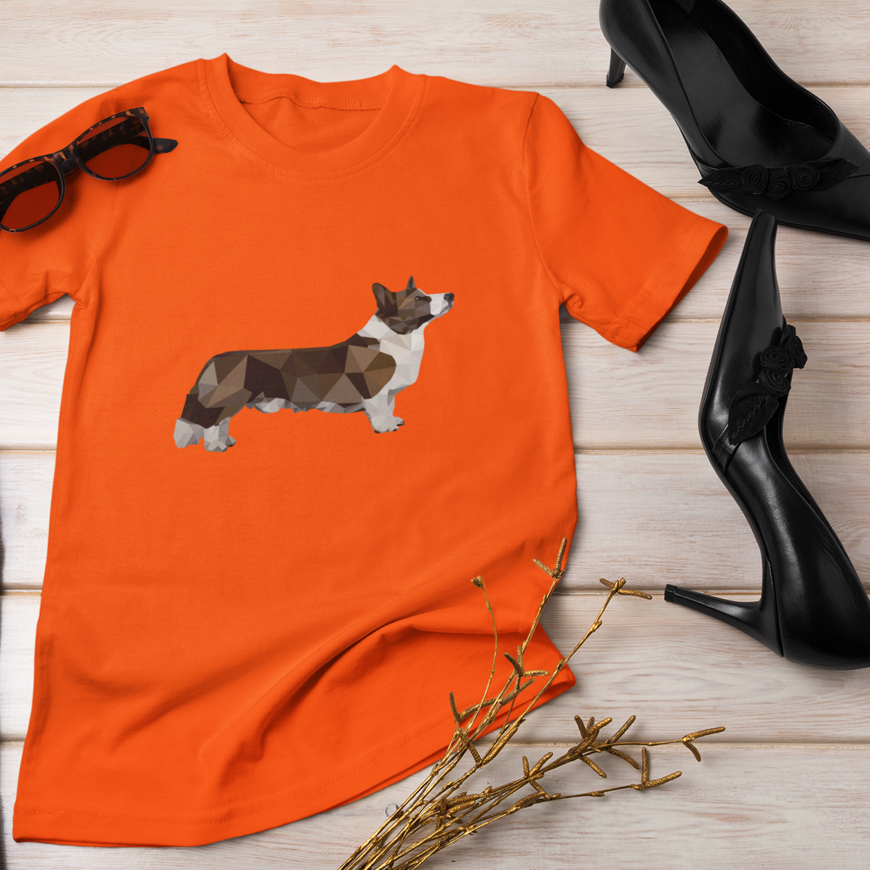 A flat lay of an orange t shirt next to some sunglasses and shoes. The design on the shirt is a polygonal cardigan corgi. 