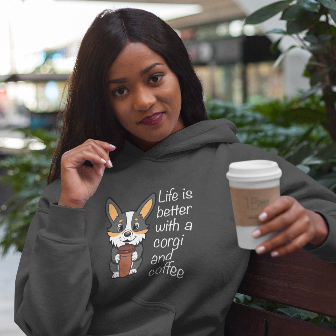 A model is drinking from a travel coffee cup and wearing wearing a hoodie with a tricolor corgi holding a cup of coffee. The text says "Life is better with a corgi and coffee". The sweatshirt is purple.