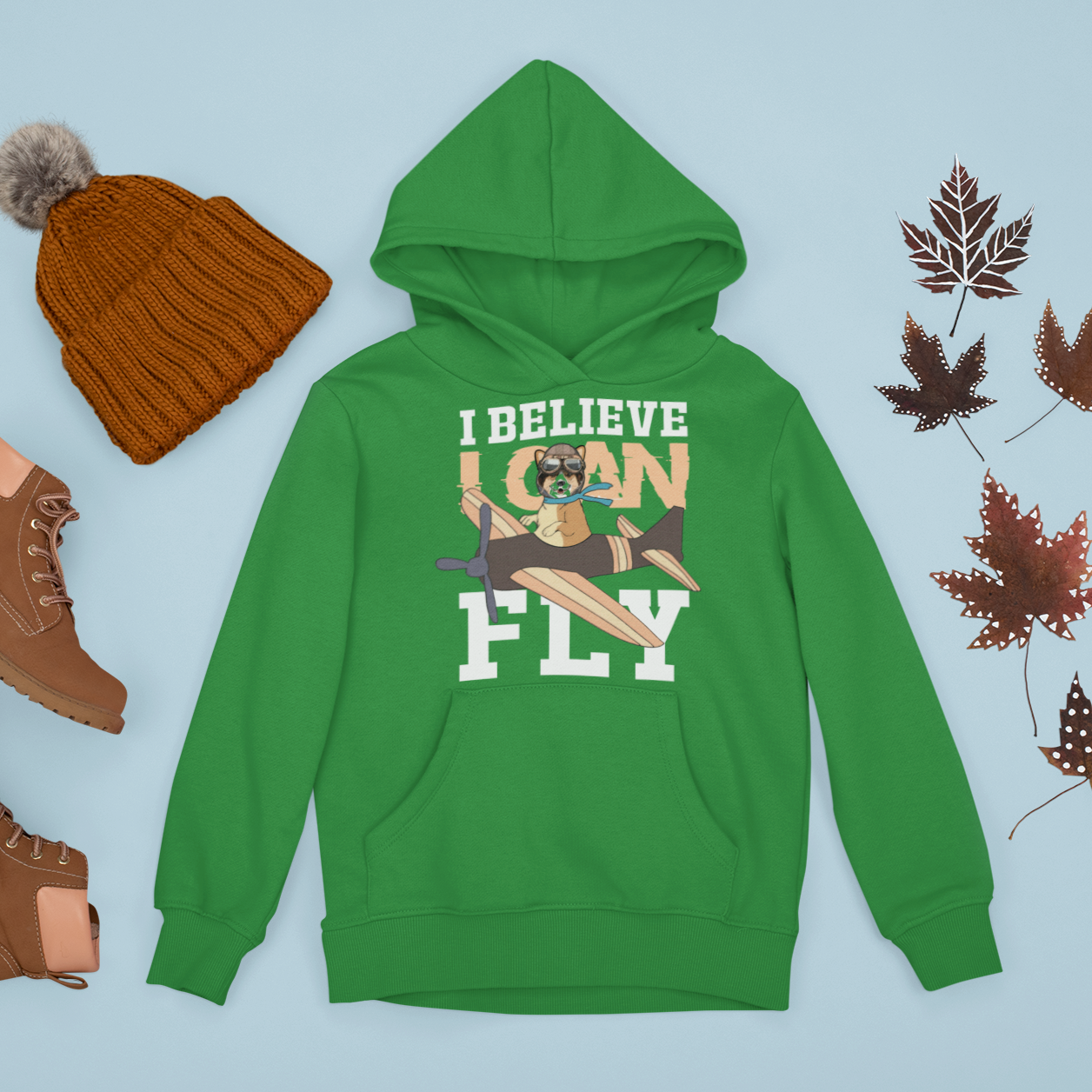 A flat lay of a green hoodie among some brown maple leaves, a knit hat, and some boots. The hoodie has a corgi flying a play with the text "I believe I can fly". 