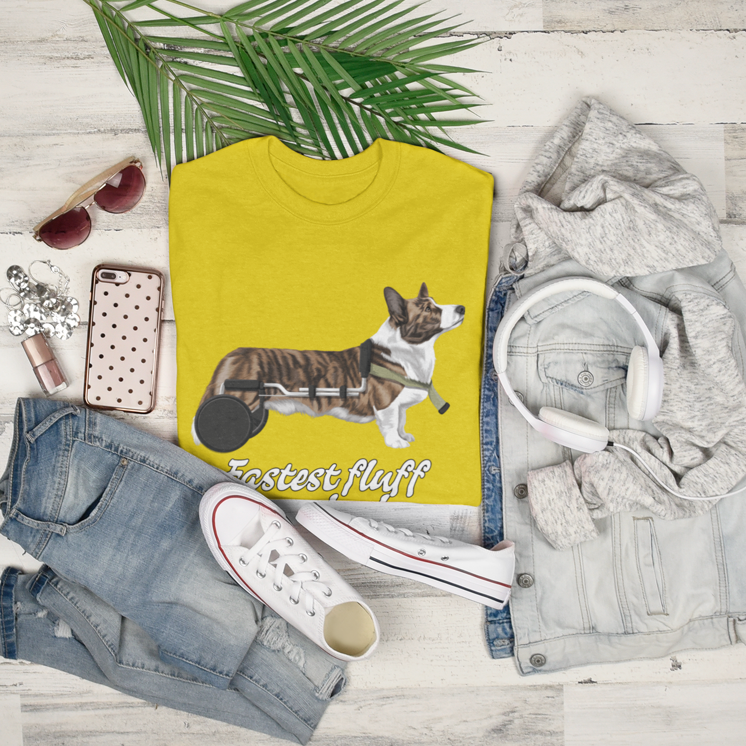 A folded t shirt among some accessories like shoes, jeans, make up, palm fronds. The design of the shirt has a cardigan corgi in a wheelchair. The text says "Fastest fluff on wheels". The shirt is daisy colored.