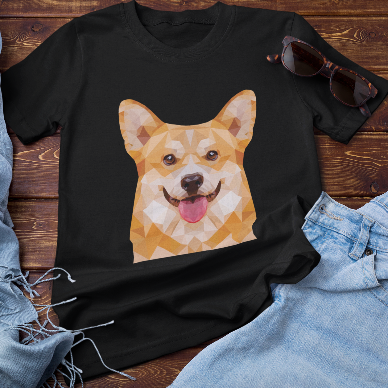 A flat lay of a t shirt next to a pair of jeans, some sunglasses, and a scarf. The t shirt is black and features a polygonal pembroke corgi. 