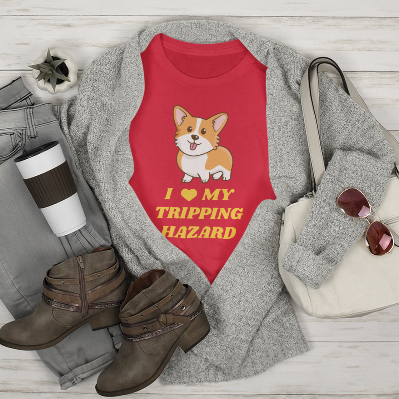 A t shirt inside an open cardigan among a pair of boots, pants, a purse, and some sunglasses. The shirt is red and has a corgi with its tongue sticking out. The text on the shirt is yellow and says "I love my tripping hazard".