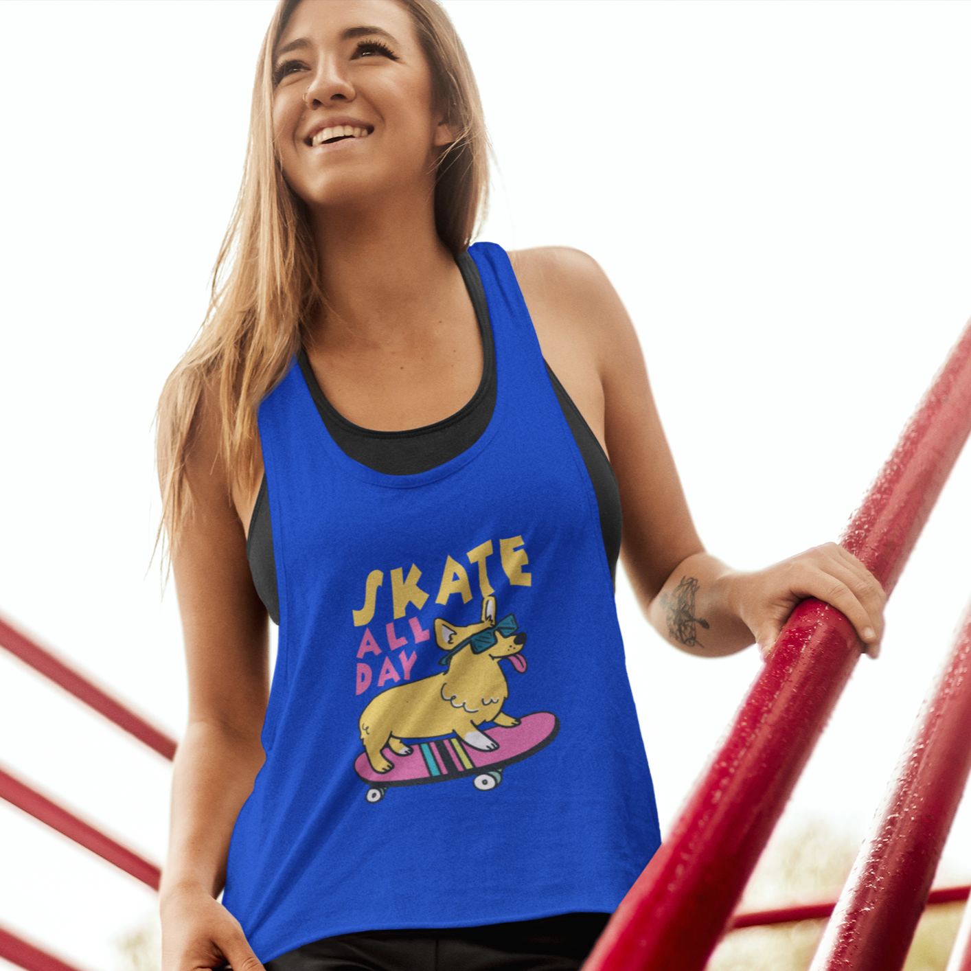 A model standing on a set of stairs wearing a racerback tank top that is blue. There is yellow and pink text that says "Skate all day". Underneath the text is a corgi wearing sunglasses, riding a skateboard. 