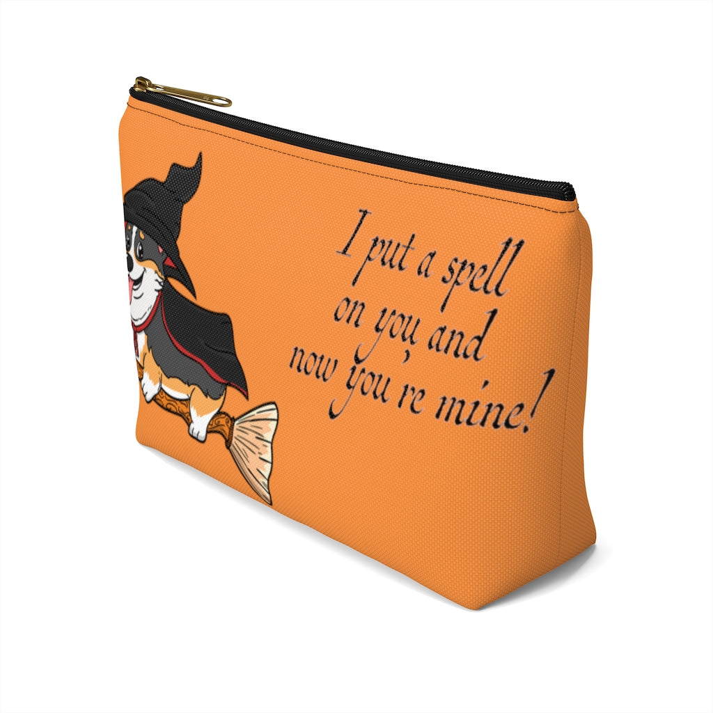 An orange make up bag with a pattern of a tricolor corgi dressed in a witch's halloween costume, on a broom. With the text "I put a spell on you and now you're mine".