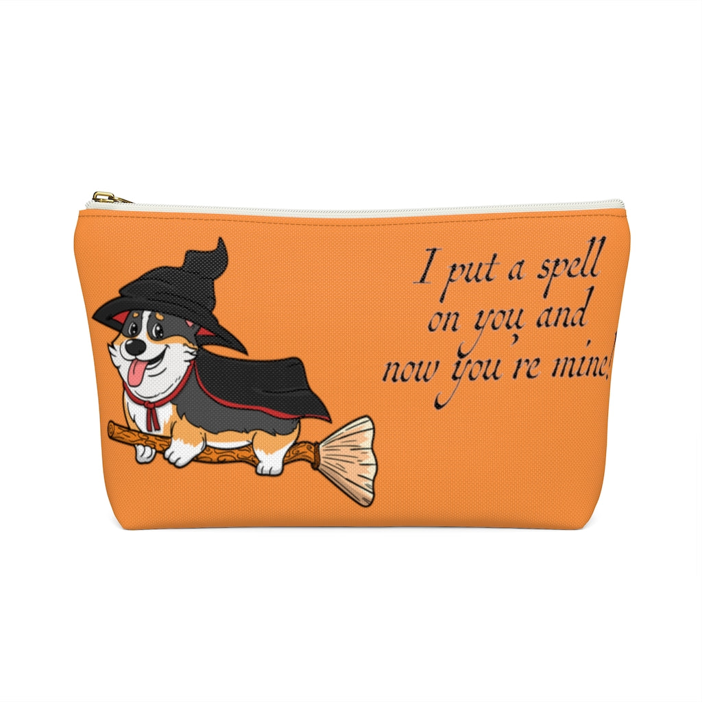 An orange make up bag with a pattern of a tricolor corgi dressed in a witch's halloween costume, on a broom. With the text "I put a spell on you and now you're mine". This picture shows the bag with a white zipper.