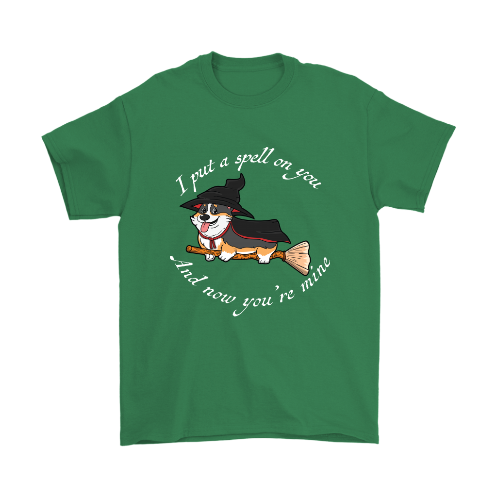 A t shirt with a tricolor corgi wearing a witch costume while riding a broom. The text says "I put a spell on you and now you're mine" and the shirt is green.