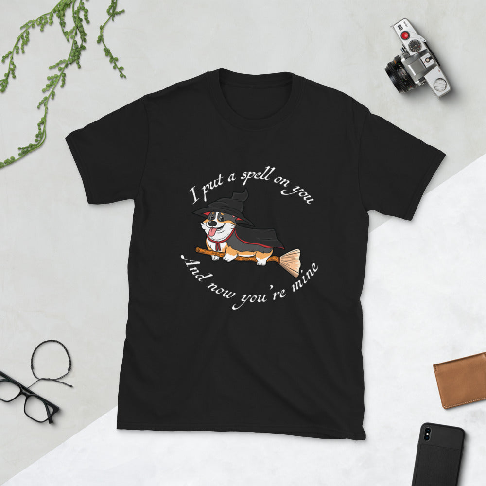 A t shirt with a tricolor corgi wearing a witch costume while riding a broom. The text says "I put a spell on you and now you're mine" and the shirt is black.