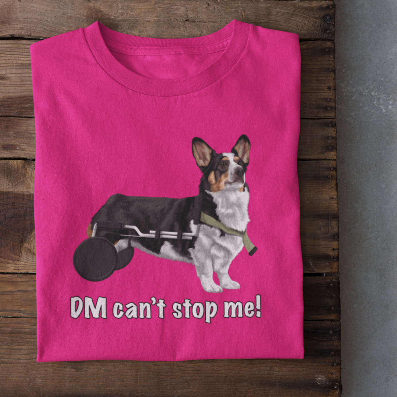 A folded, pink t shirt laying on a wooden table. The design of the shirt is a tricolor corgi using a dog wheelchair. There's white text on the shirt that says "DM can't stop me".  