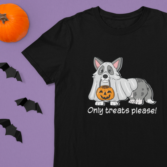 A flat lay of a short sleeved t shirt on a table with some halloween decorations. The design on the shirt shows a cardigan corgi dressed up as a ghost. The corgi has a pumpkin-shaped trick or treat bucket hanging from it's mouth. The text says "Trick or Treat!" The shirt is black.