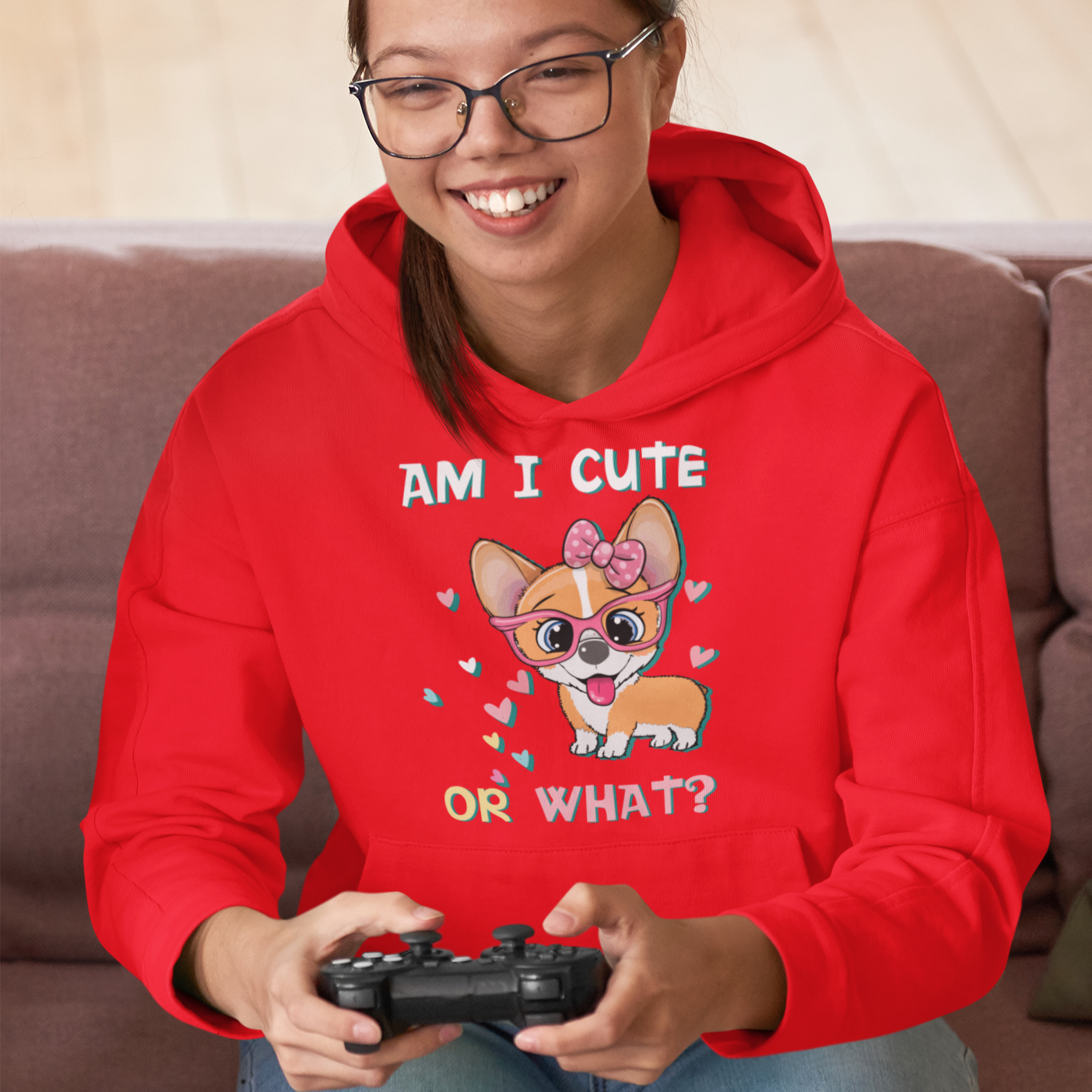 A model wearing a hoodie with the text "Am I cute or What?" with a picture of an adorable, smiling corgi. The hoodie is red. 