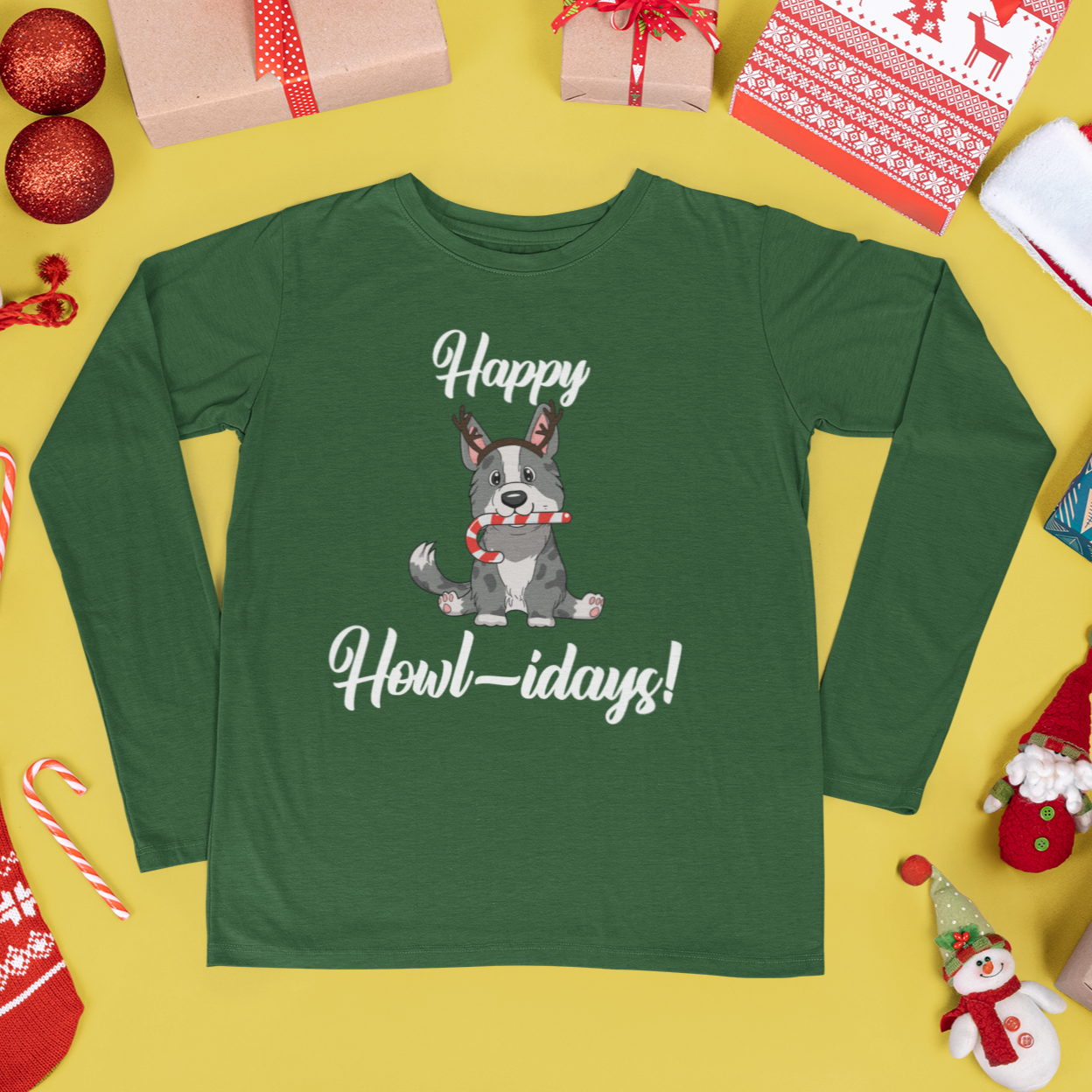 A green long sleeved shirt that has a cardigan corgi on it. The corgi is wearing fake reindeer horns and is holding a candy cane in its mouth. The text says "Happy Howl-idays". The shirt is laying among Christmas decor and presents.