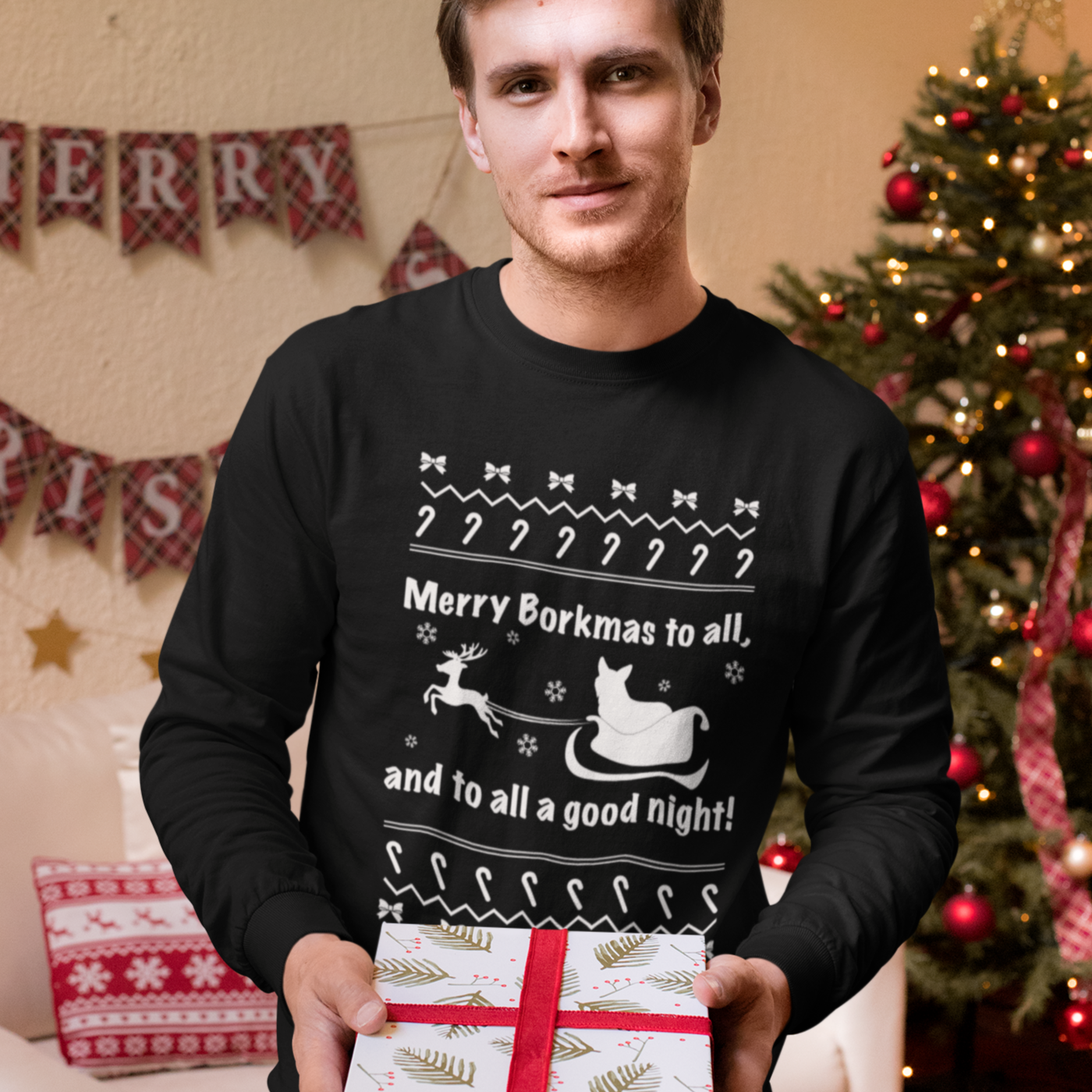 A model wearing a black long sleeved shirt with the text "Merry Borkmas to all, and to all a good night!" with a corgi in Santa's sled. He's in front of a room with Christmas decorations and he's holding a present. 