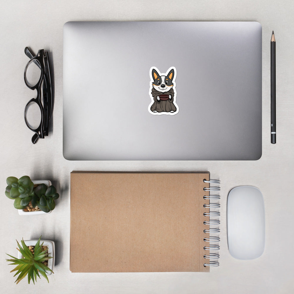 A closed laptop sitting on a desk with a sticker on it. The die cut sticker is of a corgi dressed as baby yoda holding a bowl. 