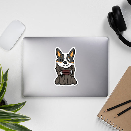 A closed laptop sitting on a desk with a sticker on it. The die cut sticker is of a corgi dressed as baby yoda holding a bowl. 