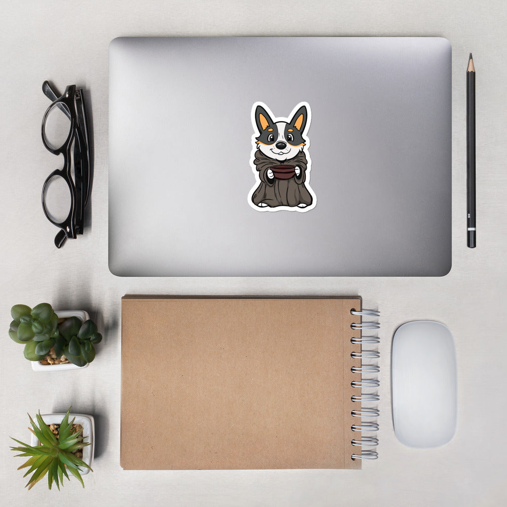 A closed laptop sitting on a desk with a sticker on it. The die cut sticker is of a corgi dressed as baby yoda holding a bowl. 