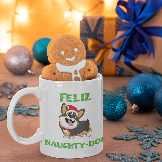 A mug that says "Feliz Naught-Dog" and has a smiling tricolor corgi wearing a Santa hat on it. The mug has a gingerbread cookie in it and is amongst Christmas decor and presents. 