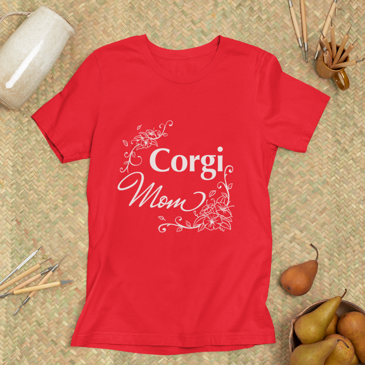 A flat lay of a red t shirt with text that says "Corgi Mom" with flowery embellishments. The background has some art supplies, a bowl of pears, and a large mug.  