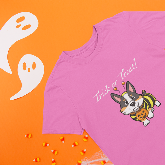 A flat lay of a short sleeved t shirt on a table with some halloween decorations. The design on the shirt shows a tricolor corgi dressed up as a bumblebee. The corgi has a pumpkin-shaped trick or treat bucket hanging from it's mouth. The text says "Trick or Treat!" The shirt is pink.