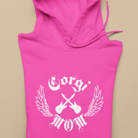 A hoodie is folded over and is laying on a table. The text on the hoodie says "Corgi Mom" in old english font. There are also crossed electric guitars and line-art wings. The hoodie is pink. 