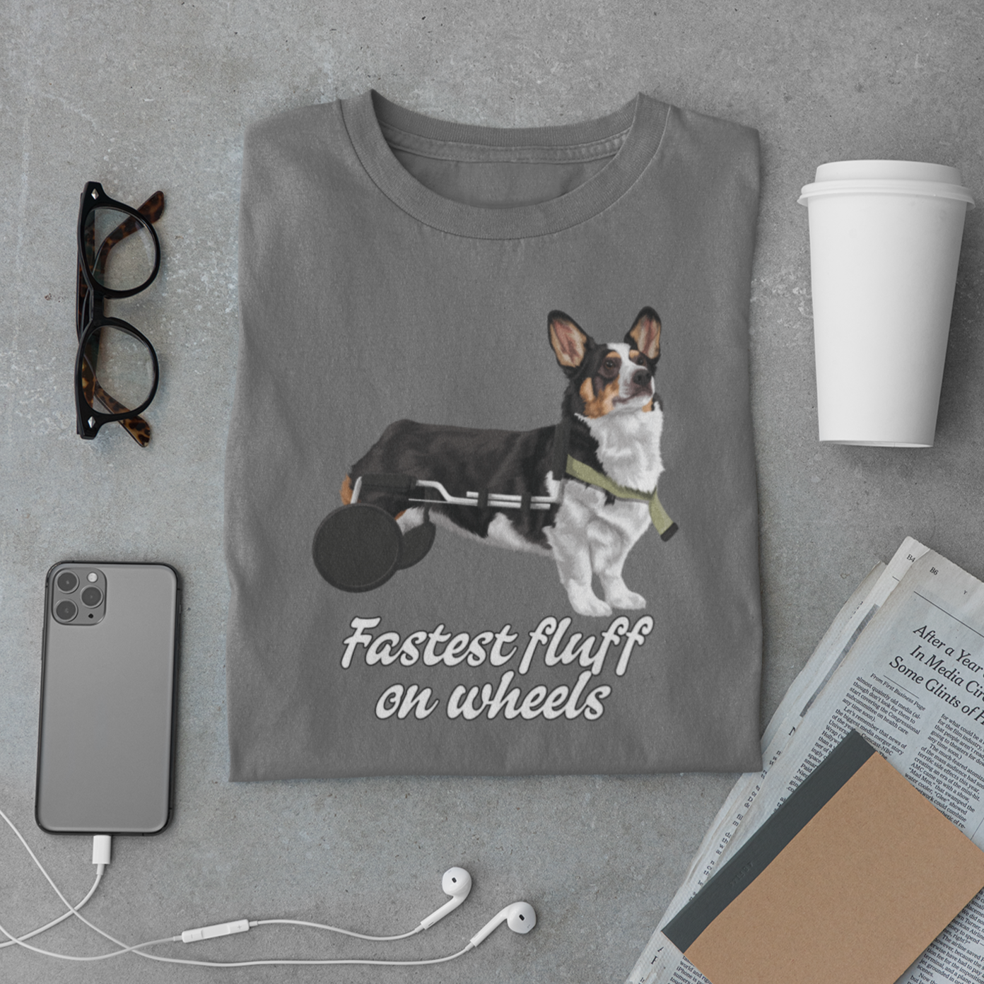 A dark grey folded t shirt among some items on a table. The design on the shirt is a tricolor corgi using a dog wheel chair. The text under the dog says Fasted fluff on wheels. 