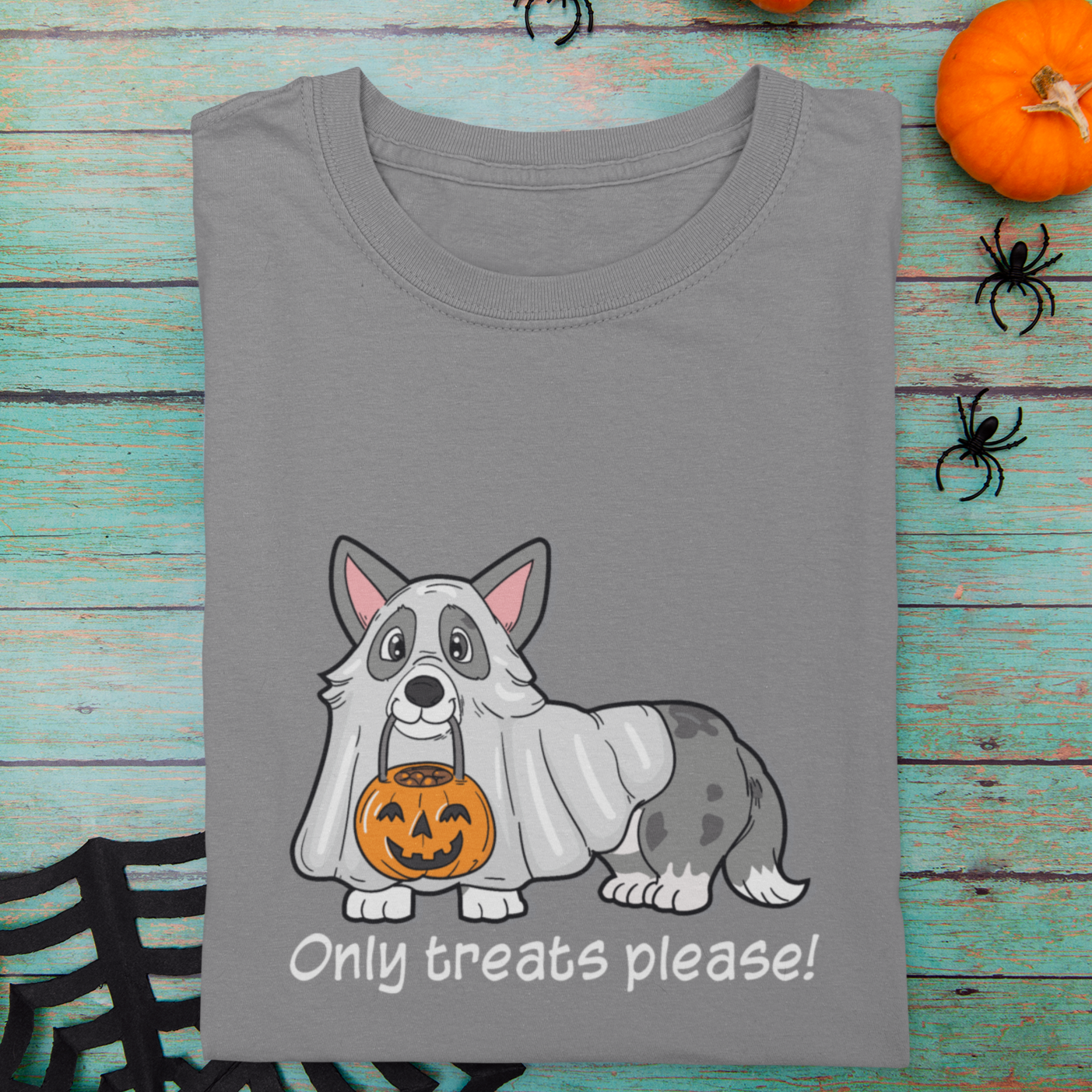 A folded t shirt on table with halloween decorations. The shirt is grey and has a cardigan corgi in a ghost costume holding a trick or treat bucket. The text says "Only treats please!"