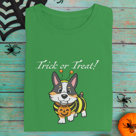 A folded long sleeved t shirt laying on a table with other halloween decoration. The  design on the shirt shows a tricolor corgi dressed up as a bumblebee. The corgi has a pumpkin-shaped trick or treat bucket hanging from it's mouth. The text says "Trick or Treat!" The shirt is green. 