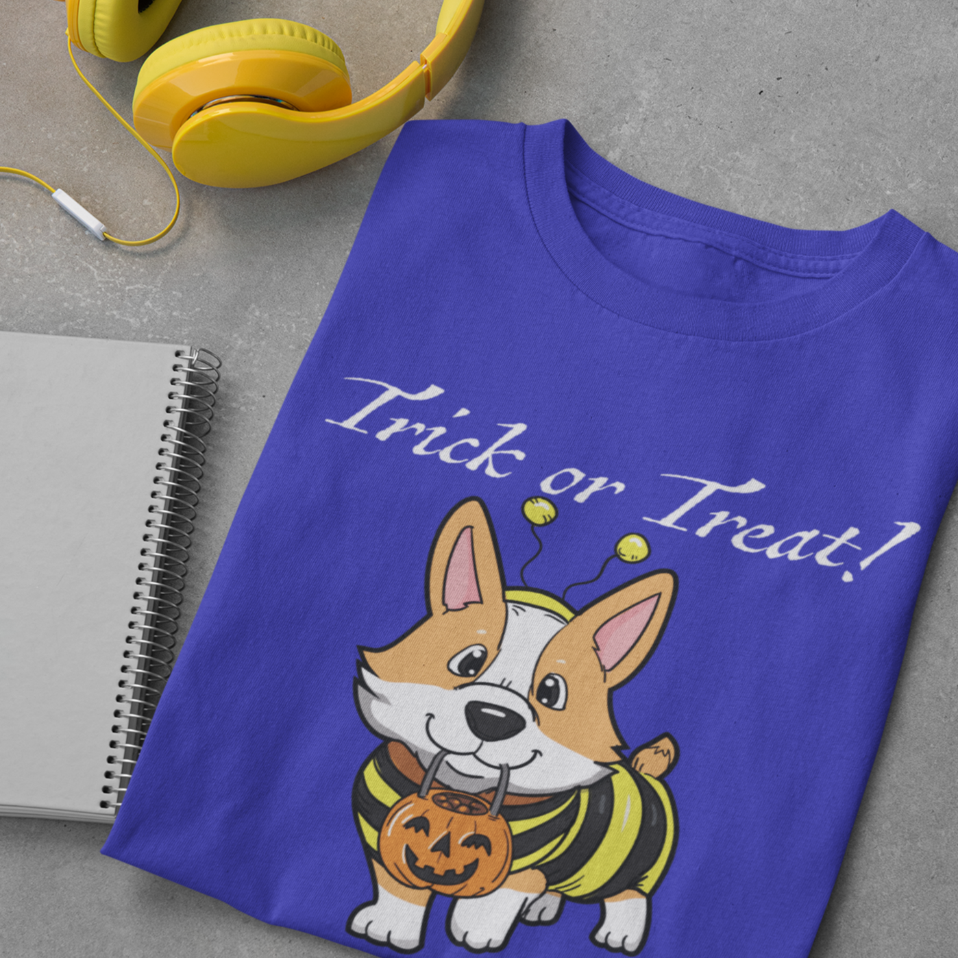 A folded t shirt is on a table with some headphones and a notebook. The design on the shirt shows a fawn corgi dressed up as a bumblebee. The corgi has a pumpkin-shaped trick or treat bucket hanging from it's mouth. The text says "Trick or Treat!" The shirt is blue.