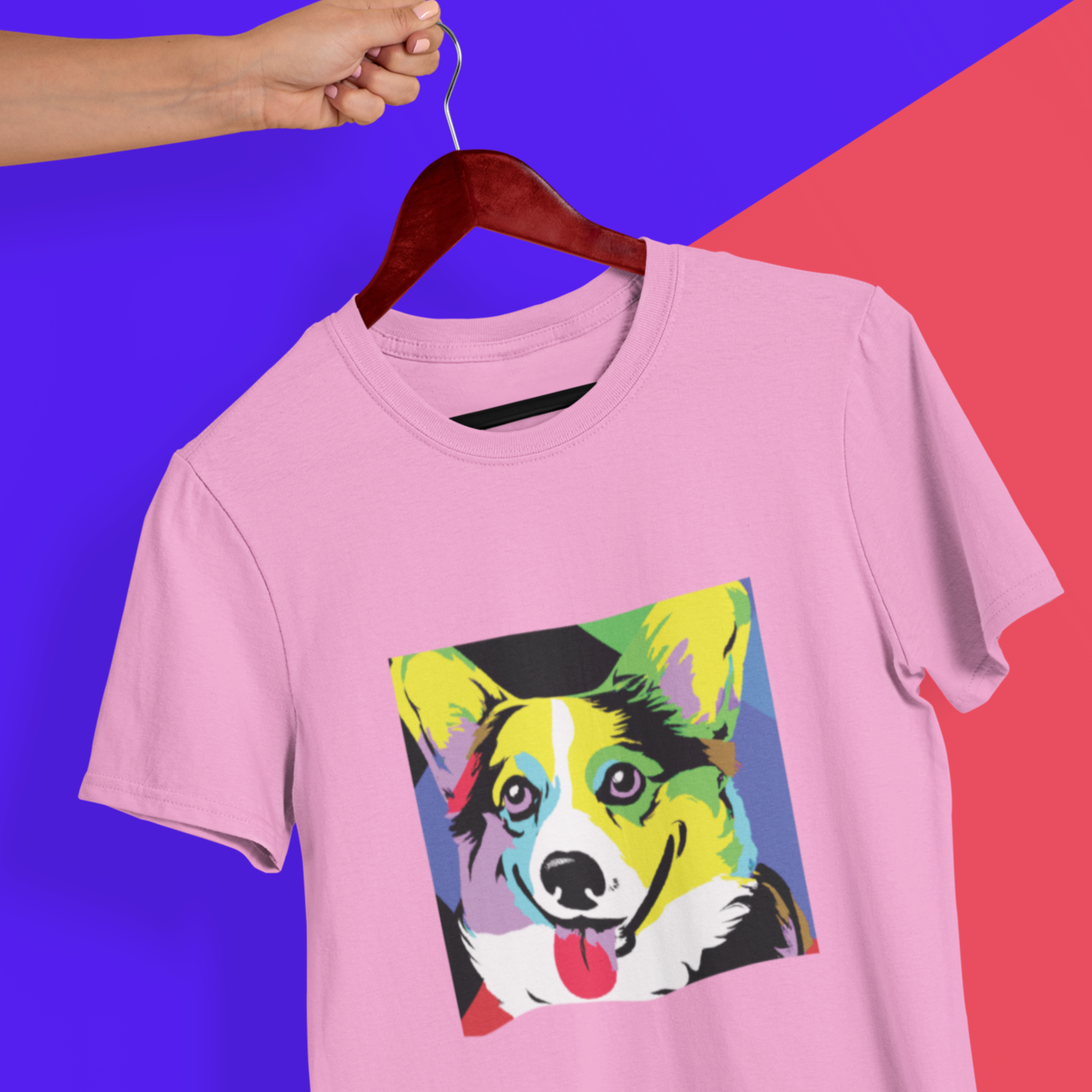 Someone is holding a wooden hanger with a t shirt hanging off of it. The pink t shirt has our Pop Art Corgi design on it. 