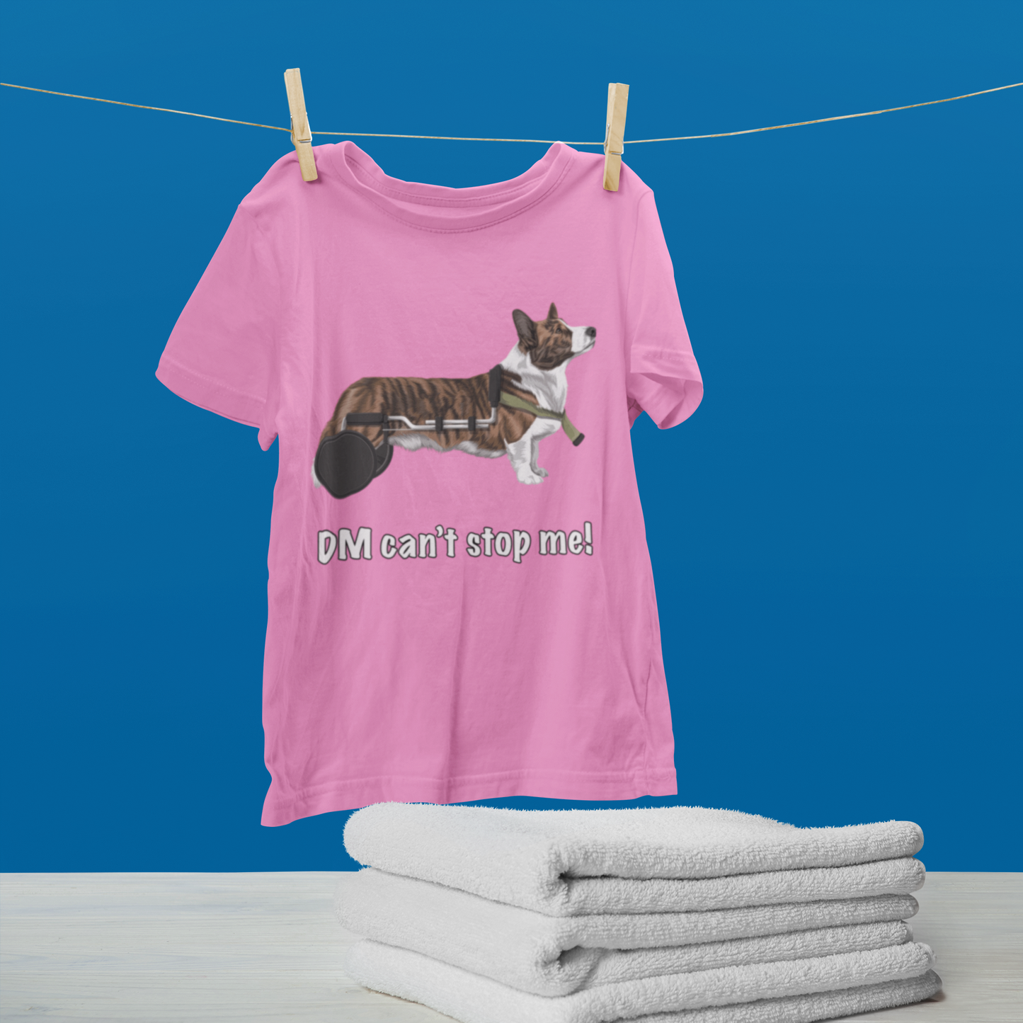 A t shirt hanging from a clothes line. There's a table below with folded towels and a blue wall in the background. The t shirt has a cardigan corgi in a wheelchair. The text says "DM can't stop me!". The shirt is pink. 