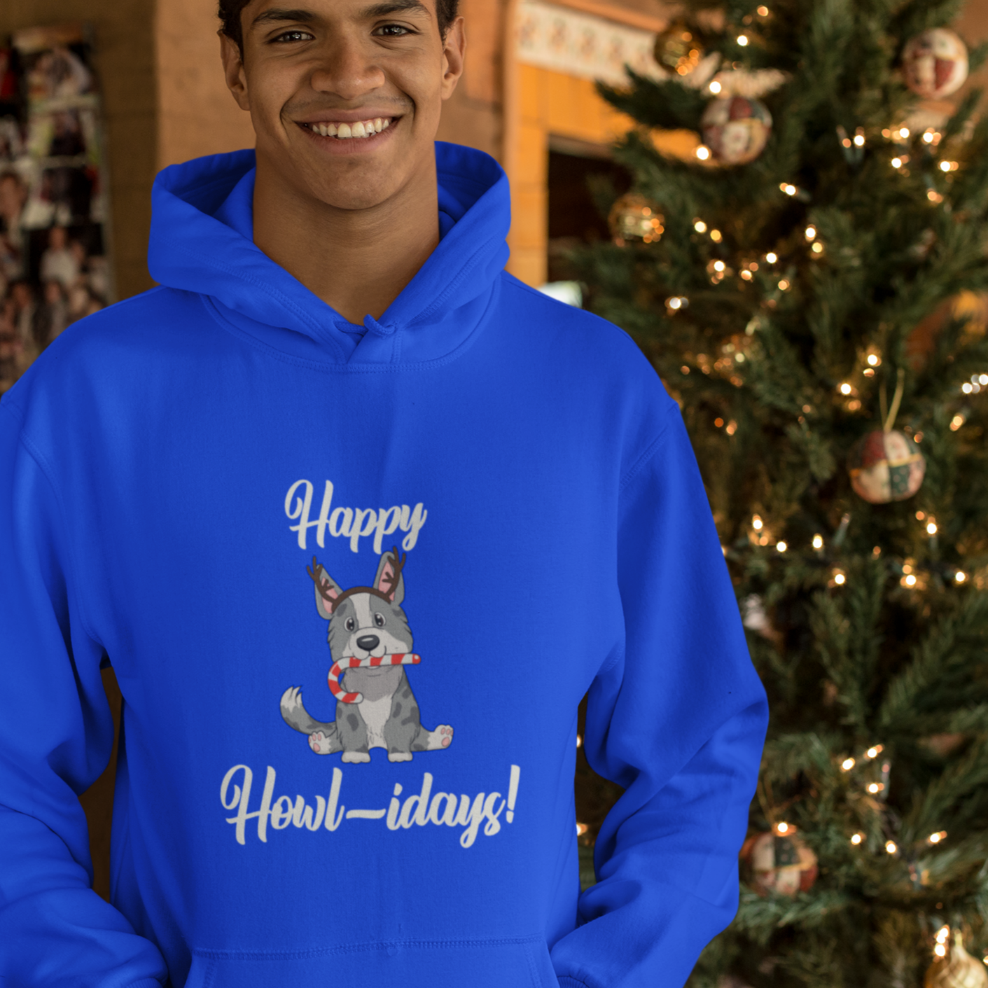 A model is wearing a blue hoodie that has a cardigan corgi on it. The corgi is wearing fake reindeer horns and is holding a candy cane in its mouth. The text says "Happy Howl-idays". The model is standing in front of a Christmas tree. 