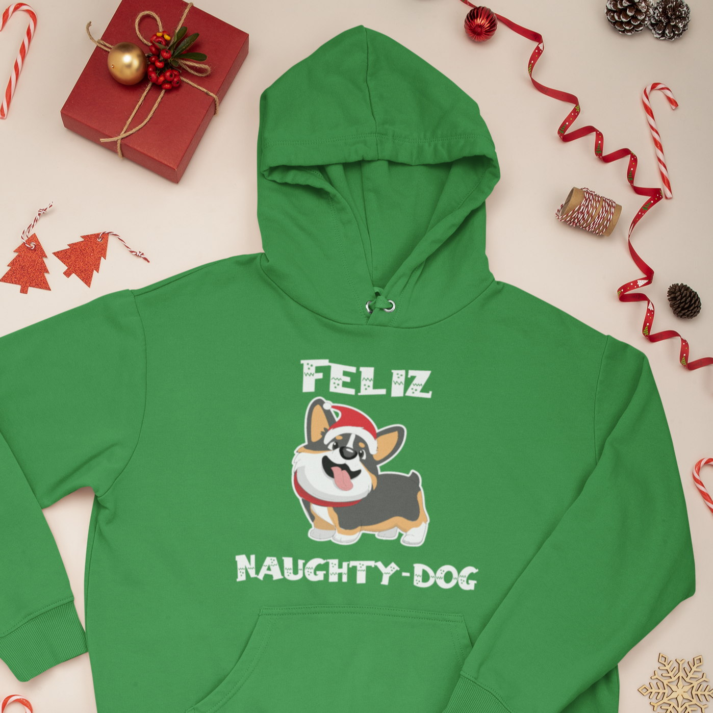 A green hoodie that says "Feliz Naught-Dog" and has a smiling tricolor corgi wearing a Santa hat on it. The hoodie is laying flat amongst Christmas decor and presents. 